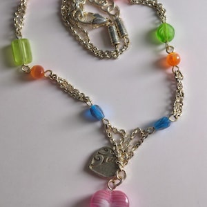 Puede incluir: Un collar de cadena plateada con una variedad de cuentas transl&uacute;cidas coloridas en tonos rosa, azul, verde y naranja. El collar incluye un dije de coraz&oacute;n plateado con un patr&oacute;n de remolinos y un colgante de coraz&oacute;n de vidrio rosa a rayas.