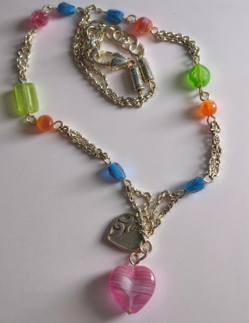 Puede incluir: Un collar de cadena plateada con una variedad de cuentas transl&uacute;cidas coloridas en tonos rosa, azul, verde y naranja. El collar incluye un dije de coraz&oacute;n plateado con un patr&oacute;n de remolinos y un colgante de coraz&oacute;n de vidrio rosa a rayas.