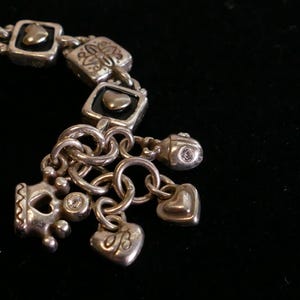 May include: Silver-toned charm bracelet with square links and various charms. Charms include hearts, crowns, and a small bead with a clear stone. The bracelet has a vintage or antique aesthetic.
