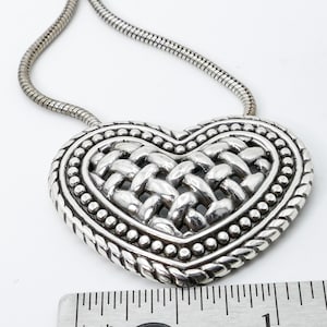 May include: Silver-toned heart pendant necklace. The heart features a woven design in the center, surrounded by a beaded border and a braided edge. The necklace has a snake chain.