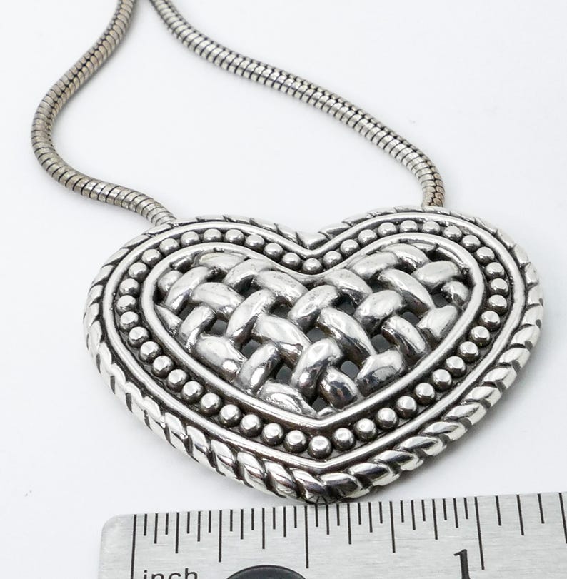 May include: Silver-toned heart pendant necklace. The heart features a woven design in the center, surrounded by a beaded border and a braided edge. The necklace has a snake chain.