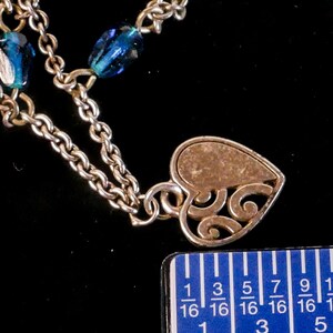 Puede incluir: Un collar de cadena de metal plateado con cuentas de vidrio azul facetado y un colgante en forma de coraz&oacute;n con un dise&ntilde;o de volutas caladas. Una regla azul est&aacute; colocada junto al colgante, mostrando una medida de unos 1,9 cent&iacute;metros.