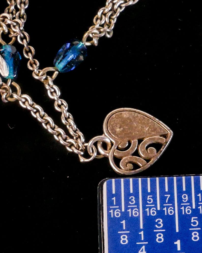 Puede incluir: Un collar de cadena de metal plateado con cuentas de vidrio azul facetado y un colgante en forma de coraz&oacute;n con un dise&ntilde;o de volutas caladas. Una regla azul est&aacute; colocada junto al colgante, mostrando una medida de unos 1,9 cent&iacute;metros.