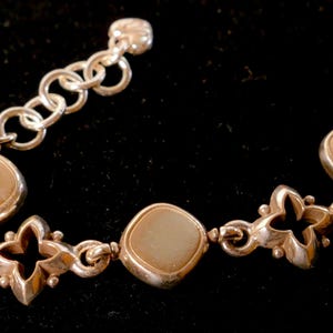 May include: Silver bracelet with alternating square and floral links. The square links have a light brown centre. The bracelet has a series of round links at the clasp. The bracelet is displayed against a black background.