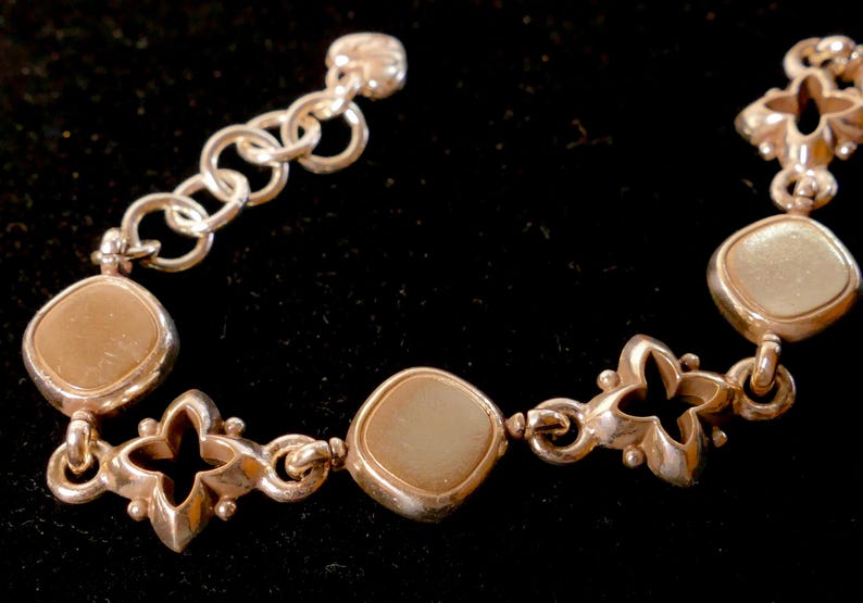May include: Silver bracelet with alternating square and floral links. The square links have a light brown centre. The bracelet has a series of round links at the clasp. The bracelet is displayed against a black background.