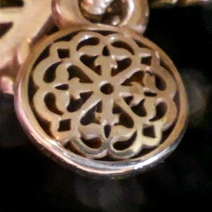 May include: A circular gold-toned metal charm featuring an intricate openwork filigree pattern with a central hole. The charm is suspended from a small circular loop at the top, showcasing a polished metallic finish and detailed cut-out design.
