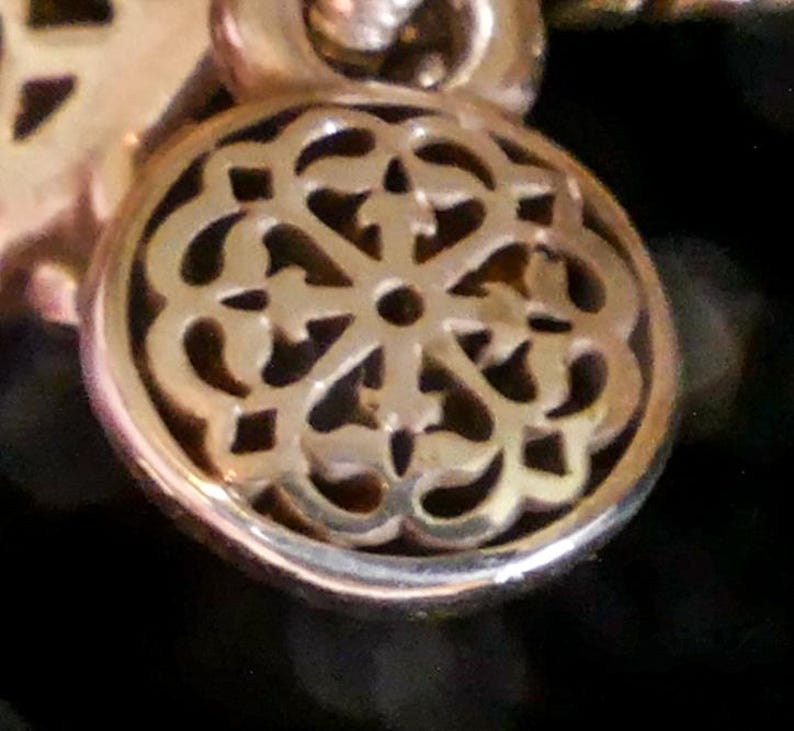 May include: A circular gold-toned metal charm featuring an intricate openwork filigree pattern with a central hole. The charm is suspended from a small circular loop at the top, showcasing a polished metallic finish and detailed cut-out design.