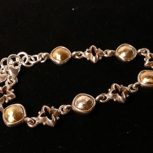 May include: Silver bracelet with gold-coloured square and round accents. The bracelet features a series of gold squares and decorative silver links, including heart and flower shapes. The bracelet is displayed against a black background.