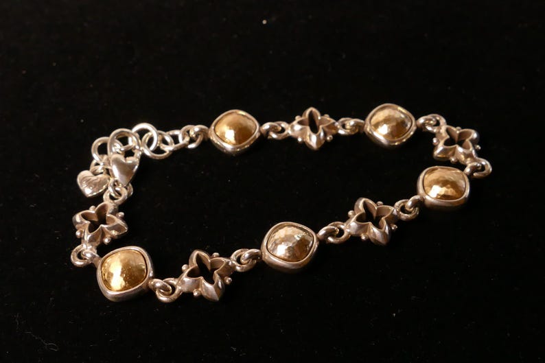 May include: Silver bracelet with gold-coloured square and round accents. The bracelet features a series of gold squares and decorative silver links, including heart and flower shapes. The bracelet is displayed against a black background.
