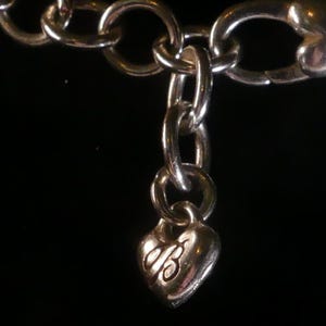 Puede incluir: Una pulsera de cadena de metal plateado con un peque&ntilde;o dije en forma de coraz&oacute;n abombado que cuelga de un eslab&oacute;n. El dije de coraz&oacute;n tiene grabada una letra B en cursiva estilizada en su superficie frontal, reflejando luz sobre un fondo oscuro.