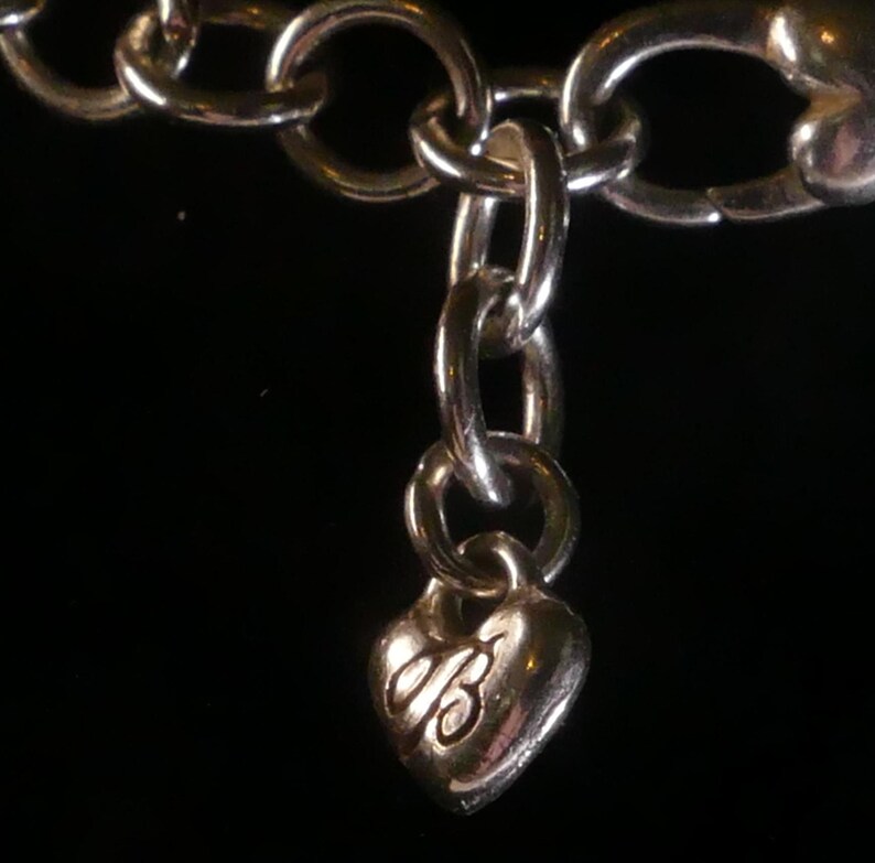 Puede incluir: Una pulsera de cadena de metal plateado con un peque&ntilde;o dije en forma de coraz&oacute;n abombado que cuelga de un eslab&oacute;n. El dije de coraz&oacute;n tiene grabada una letra B en cursiva estilizada en su superficie frontal, reflejando luz sobre un fondo oscuro.