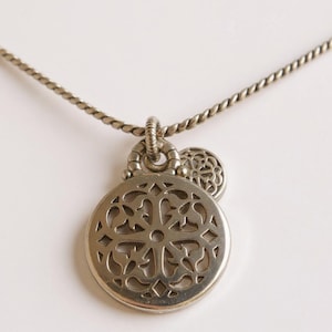 May include: A silver-toned necklace featuring a circular pendant with an intricate openwork cutout pattern. The pendant is attached to a twisted metal chain by a textured ring bail, with a smaller, matching circular charm layered behind it.