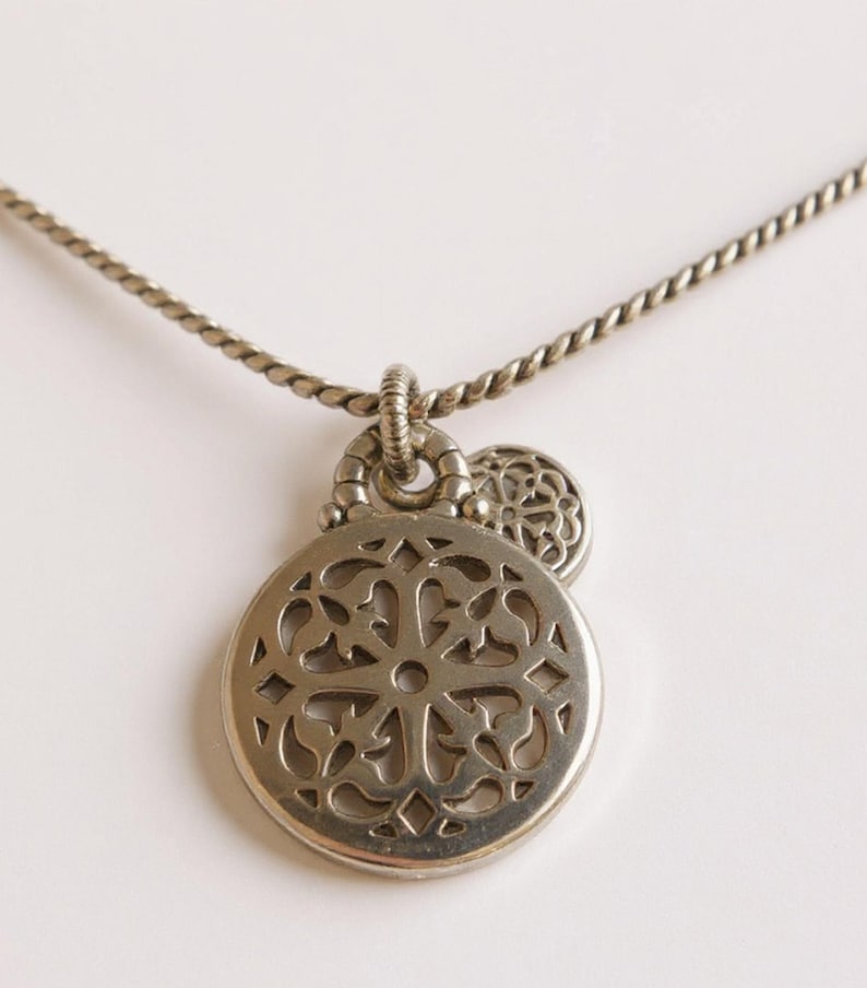 May include: A silver-toned necklace featuring a circular pendant with an intricate openwork cutout pattern. The pendant is attached to a twisted metal chain by a textured ring bail, with a smaller, matching circular charm layered behind it.