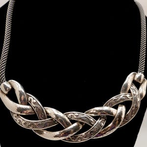 May include: Silver-toned statement necklace with a woven design. The necklace features large, hammered-look links with intricate etched patterns. The chain is a fine, woven mesh. The necklace is displayed against a black background.