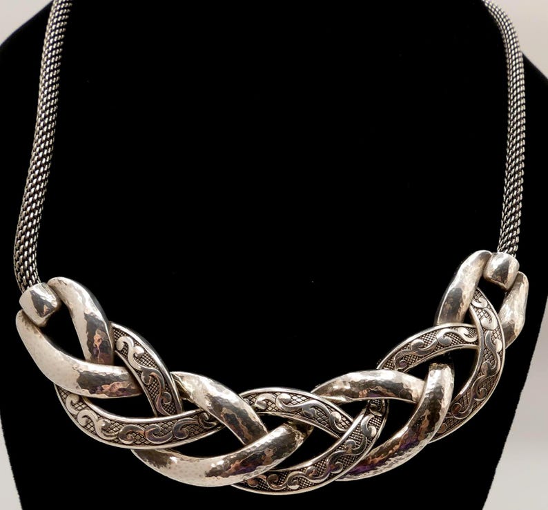 May include: Silver-toned statement necklace with a woven design. The necklace features large, hammered-look links with intricate etched patterns. The chain is a fine, woven mesh. The necklace is displayed against a black background.