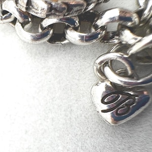 May include: Close-up of a silver-toned chain bracelet. The bracelet features a heart-shaped charm with a cursive "B" and a series of interlocking links. The bracelet is on a white surface.