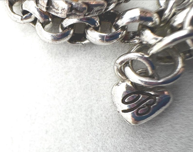 May include: Close-up of a silver-toned chain bracelet. The bracelet features a heart-shaped charm with a cursive "B" and a series of interlocking links. The bracelet is on a white surface.