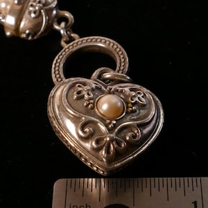May include: A silver-toned heart-shaped pendant with a pearl center and ornate floral detailing. The pendant is connected to a chain and ring. The image includes a ruler showing inches.