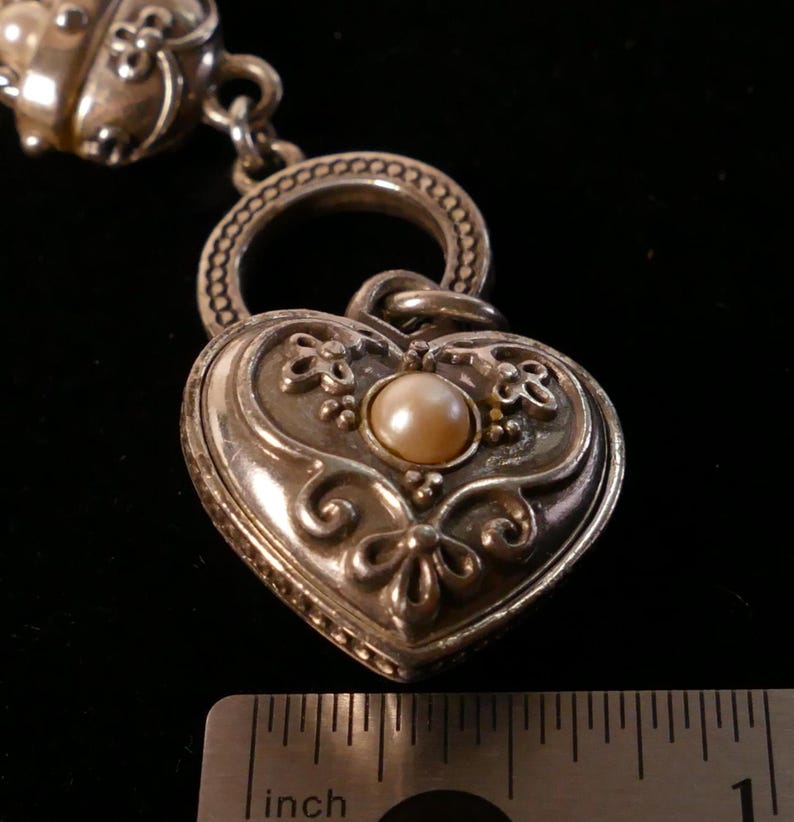 May include: A silver-toned heart-shaped pendant with a pearl center and ornate floral detailing. The pendant is connected to a chain and ring. The image includes a ruler showing inches.