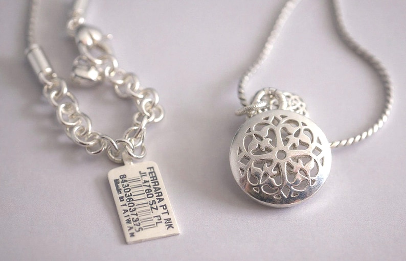 May include: A silver-toned necklace featuring a round pendant with an intricate cut-out filigree pattern. The chain includes a lobster clasp and a rectangular white tag printed with the text FERRARA PT NK JL4760 SZ PL 843036037975 Made in TAIWAN.
