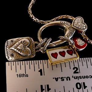 Może przedstawiać: Srebrzysty naszyjnik z dużym okrągłym zapięciem i kilkoma zawieszkami w kształcie serca, w tym jedną z czerwoną emalią i trzema małymi sercami, obok linijki pokazującej, że zawieszki mają około 3,8 cm szerokości.