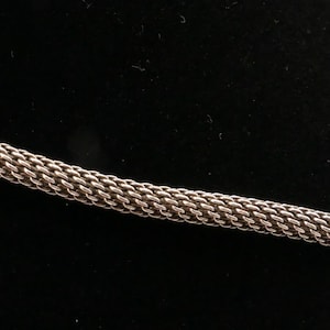 May include: Close-up of a silver-colored chain with a woven design. The chain is made of small, interlocking links, creating a textured surface. The background is a solid black.