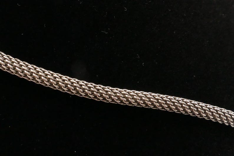 May include: Close-up of a silver-colored chain with a woven design. The chain is made of small, interlocking links, creating a textured surface. The background is a solid black.