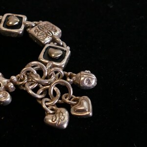 May include: Silver-toned bracelet with square links, heart-shaped charms, and a small charm with a single clear stone. The bracelet features decorative floral and heart motifs, and is set against a black background.