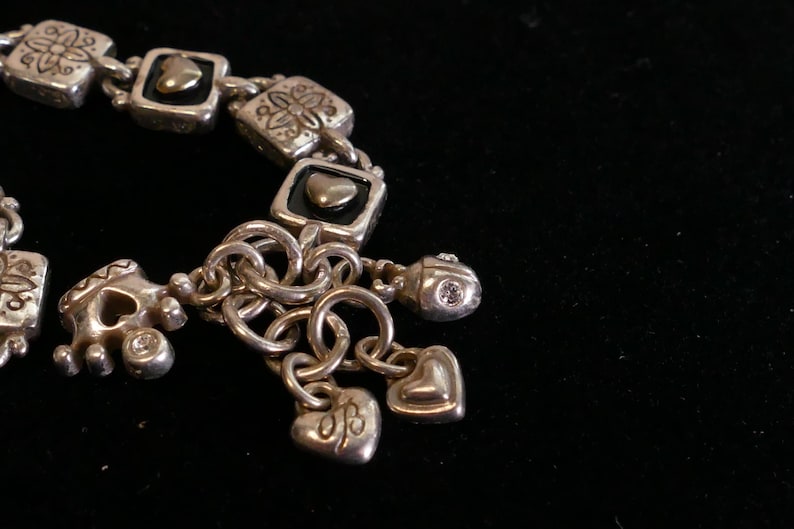 May include: Silver-toned bracelet with square links, heart-shaped charms, and a small charm with a single clear stone. The bracelet features decorative floral and heart motifs, and is set against a black background.