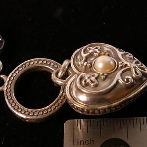 May include: Close-up of a silver heart-shaped pendant with a pearl center and ornate floral detailing. The pendant is attached to a silver ring and a braided strap. A ruler shows the pendant is approximately 1 inch in length.