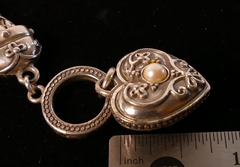 May include: Close-up of a silver heart-shaped pendant with a pearl center and ornate floral detailing. The pendant is attached to a silver ring and a braided strap. A ruler shows the pendant is approximately 1 inch in length.