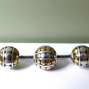 K&ouml;nnte beinhalten: Eine Kette mit dekorativen Perlen mit einem silbernen und goldenen Punktmuster. Die Perlen sind kugelf&ouml;rmig und auf einer silbernen Kette aufgereiht. Der Hintergrund ist ein weiches Gr&uuml;n und Wei&szlig;.