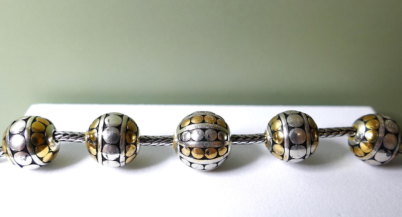 K&ouml;nnte beinhalten: Eine Kette mit dekorativen Perlen mit einem silbernen und goldenen Punktmuster. Die Perlen sind kugelf&ouml;rmig und auf einer silbernen Kette aufgereiht. Der Hintergrund ist ein weiches Gr&uuml;n und Wei&szlig;.