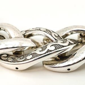 May include: Close-up of a silver chain necklace with a hammered texture. The links are large and oval-shaped, with decorative scrollwork. The necklace is displayed against a white background.