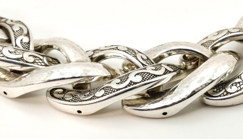 May include: Close-up of a silver chain necklace with a hammered texture. The links are large and oval-shaped, with decorative scrollwork. The necklace is displayed against a white background.