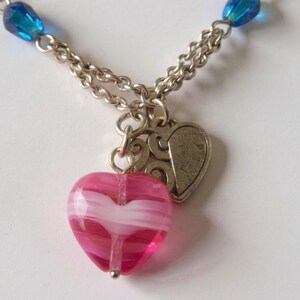 Puede incluir: Un collar con cadena de tono plateado, cuentas de vidrio azul y un colgante central que consiste en un dije de coraz&oacute;n de vidrio veteado en rosa y blanco junto a un dije de coraz&oacute;n de metal plateado texturizado con dise&ntilde;o en relieve.