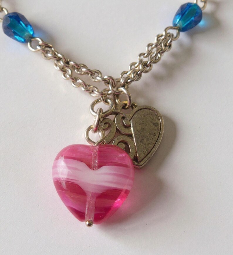 Puede incluir: Un collar con cadena de tono plateado, cuentas de vidrio azul y un colgante central que consiste en un dije de coraz&oacute;n de vidrio veteado en rosa y blanco junto a un dije de coraz&oacute;n de metal plateado texturizado con dise&ntilde;o en relieve.