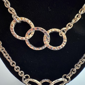 Puede incluir: Collar plateado con dise&ntilde;o de cadena y tres anillos texturizados entrelazados. El collar se exhibe sobre un fondo negro, resaltando los intrincados detalles de la joya. Los eslabones de la cadena son claramente visibles.