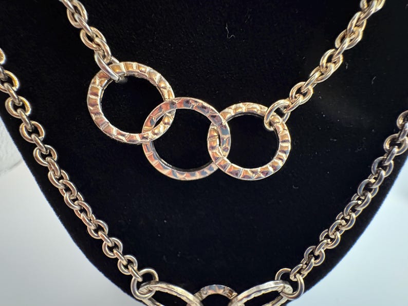 Puede incluir: Collar plateado con dise&ntilde;o de cadena y tres anillos texturizados entrelazados. El collar se exhibe sobre un fondo negro, resaltando los intrincados detalles de la joya. Los eslabones de la cadena son claramente visibles.
