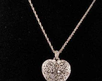 May include: A silver-toned heart-shaped pendant necklace featuring an intricate filigree pattern with embedded clear stones. The pendant hangs from a delicate twisted rope chain, creating a cohesive piece of jewelry with a metallic finish.