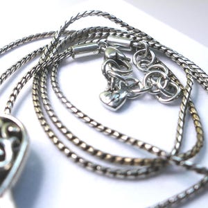 Puede incluir: Primer plano de un collar plateado con un dise&ntilde;o de cadena detallado. El collar presenta un cierre en forma de coraz&oacute;n y un colgante decorativo. El metal tiene un acabado brillante y el dise&ntilde;o es intrincado.