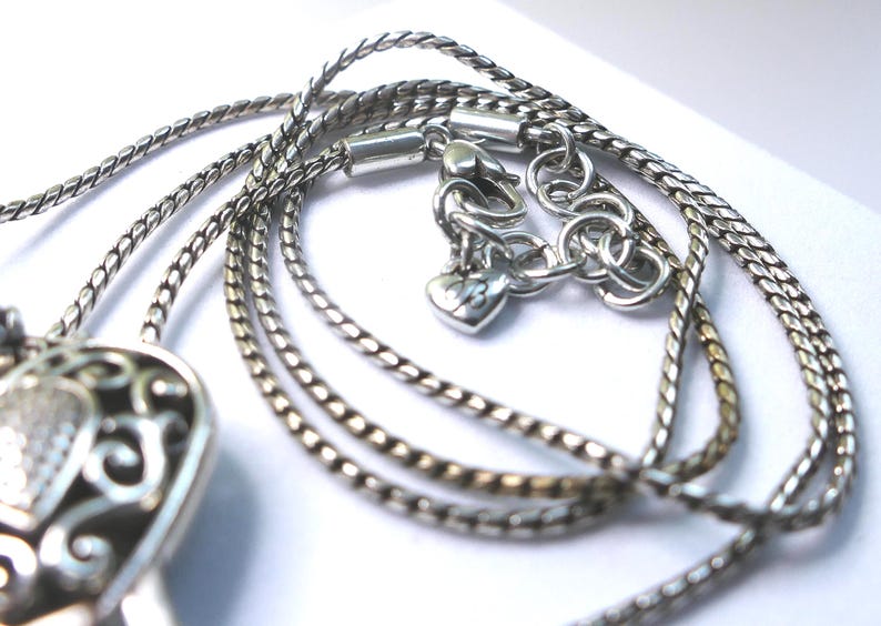 Puede incluir: Primer plano de un collar plateado con un dise&ntilde;o de cadena detallado. El collar presenta un cierre en forma de coraz&oacute;n y un colgante decorativo. El metal tiene un acabado brillante y el dise&ntilde;o es intrincado.