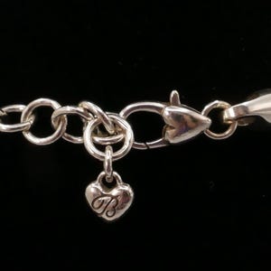 May include: Silver-toned necklace clasp and chain with a heart-shaped charm. The charm has the letter "B" engraved on it. The chain is made of small, interconnected rings. The background is black.