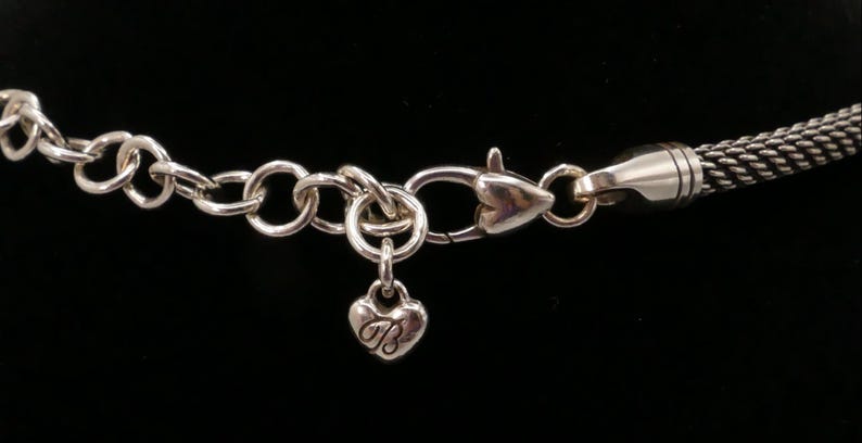 May include: Silver-toned necklace clasp and chain with a heart-shaped charm. The charm has the letter "B" engraved on it. The chain is made of small, interconnected rings. The background is black.