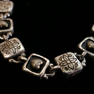 May include: Silver-toned bracelet with square links featuring floral and heart-shaped accents. The bracelet has a vintage aesthetic, with a dark background that highlights the intricate details of the jewelry.