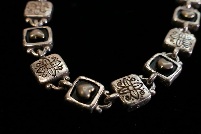 May include: Silver-toned bracelet with square links featuring floral and heart-shaped accents. The bracelet has a vintage aesthetic, with a dark background that highlights the intricate details of the jewelry.