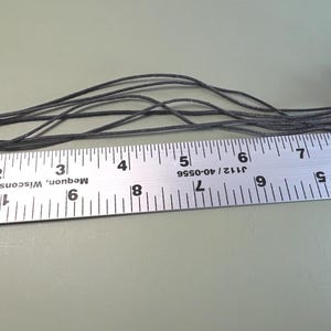 May include: A multi-strand black cord necklace featuring silver-toned heart-shaped charms with embossed patterns. The necklace is positioned alongside a metal ruler, showing a total length of approximately 11 inches from end to end.