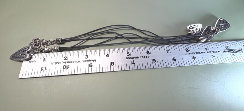 May include: A multi-strand black cord necklace featuring silver-toned heart-shaped charms with embossed patterns. The necklace is positioned alongside a metal ruler, showing a total length of approximately 11 inches from end to end.