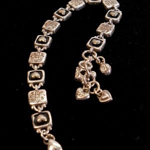 May include: Silver-toned bracelet with square links, each featuring a heart and floral design. The bracelet has dangling charms, including hearts and a small, jeweled bead. A lobster clasp secures the bracelet.