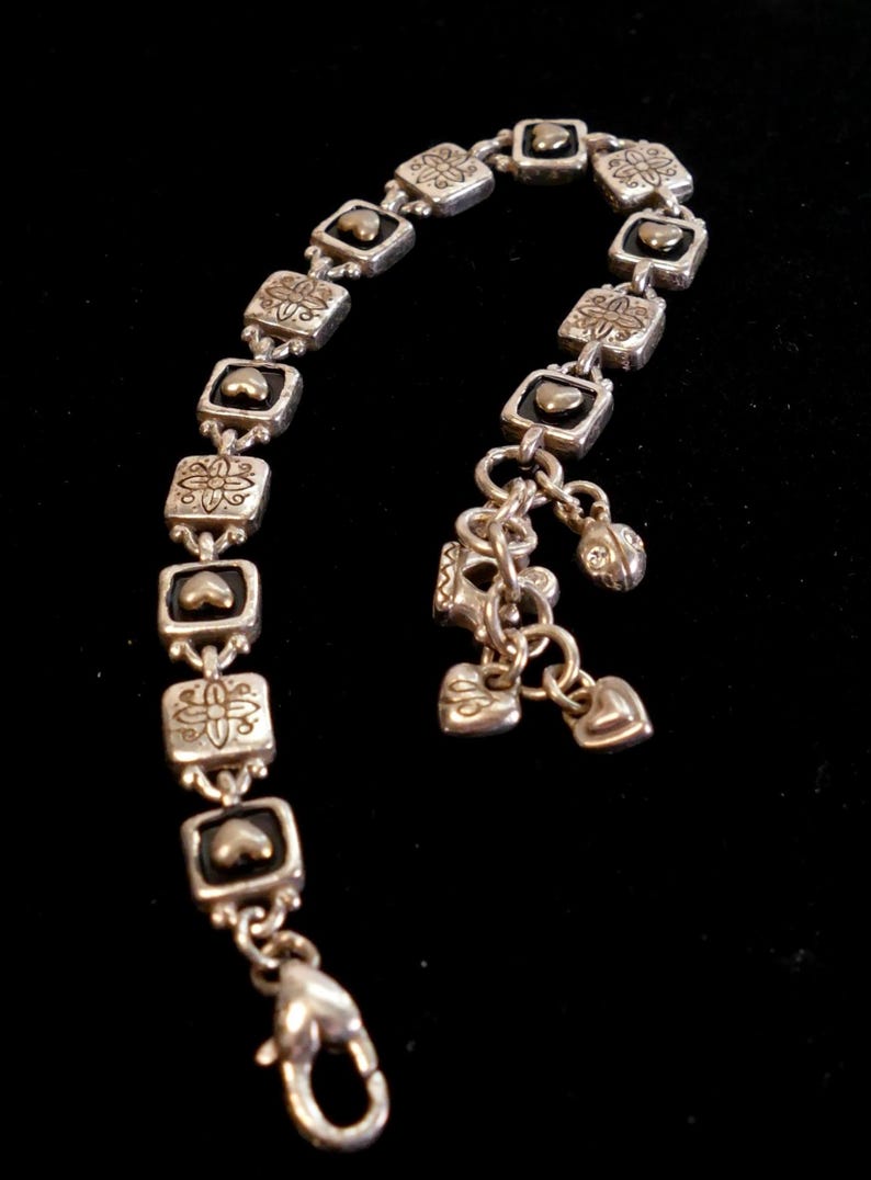 May include: Silver-toned bracelet with square links, each featuring a heart and floral design. The bracelet has dangling charms, including hearts and a small, jeweled bead. A lobster clasp secures the bracelet.